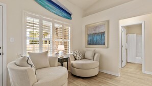 A living room with two beige armchairs, a side table with a lamp, a painting on the wall, and a view through a window with blinds. - Here Comes Sunshine by Southern Vacation Rentals (Miramar Beach)