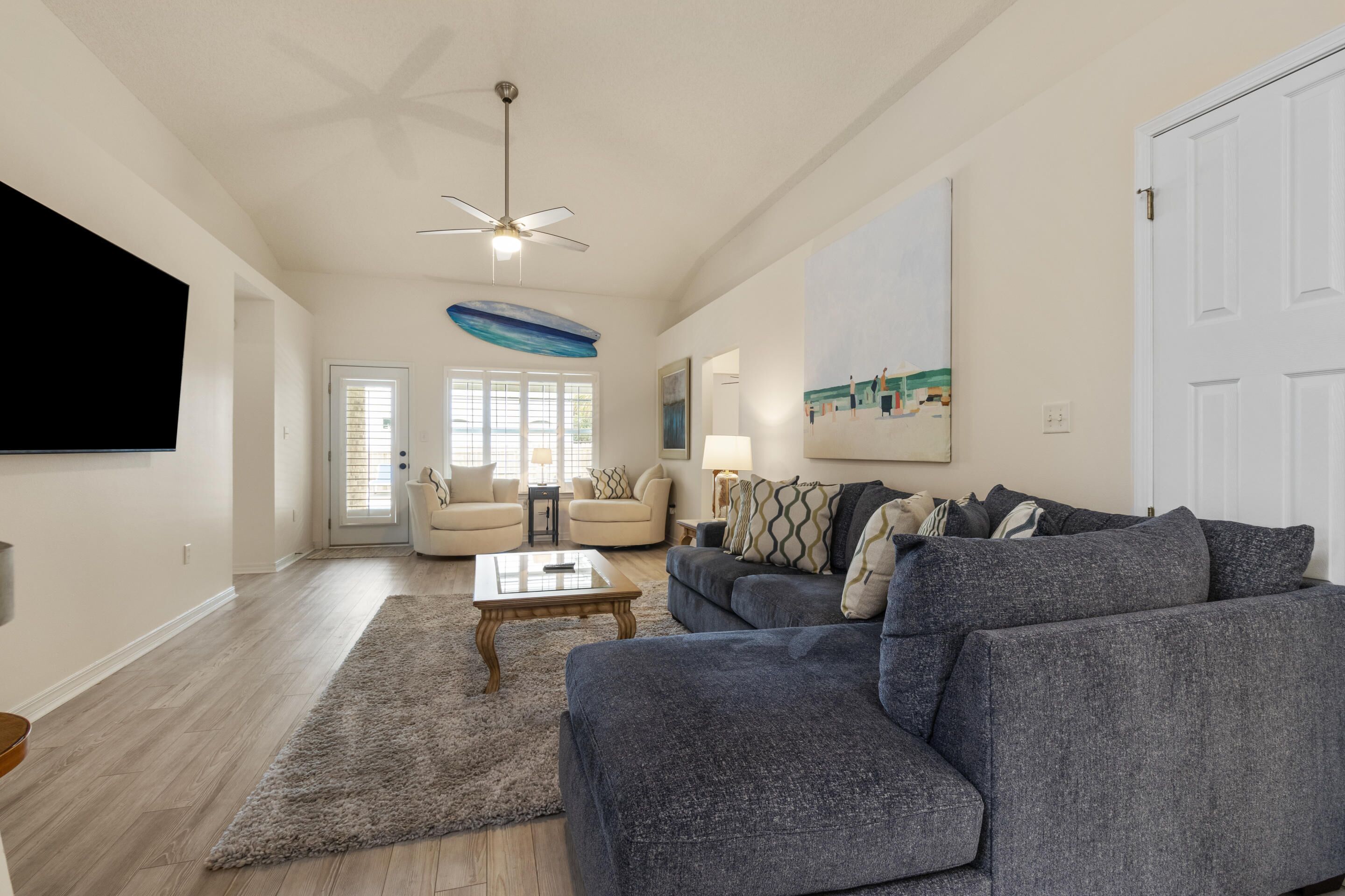 Un salon spacieux avec un grand canapé d’angle gris, une table basse et un ventilateur de plafond. Une porte donne accès à une autre pièce, et un tableau est accroché au mur.