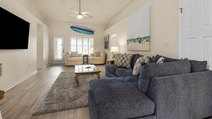 A spacious living room with a large gray sectional sofa, a coffee table, and a ceiling fan. There is a door leading to another room, and a painting on the wall. - Here Comes Sunshine by Southern Vacation Rentals (Miramar Beach)