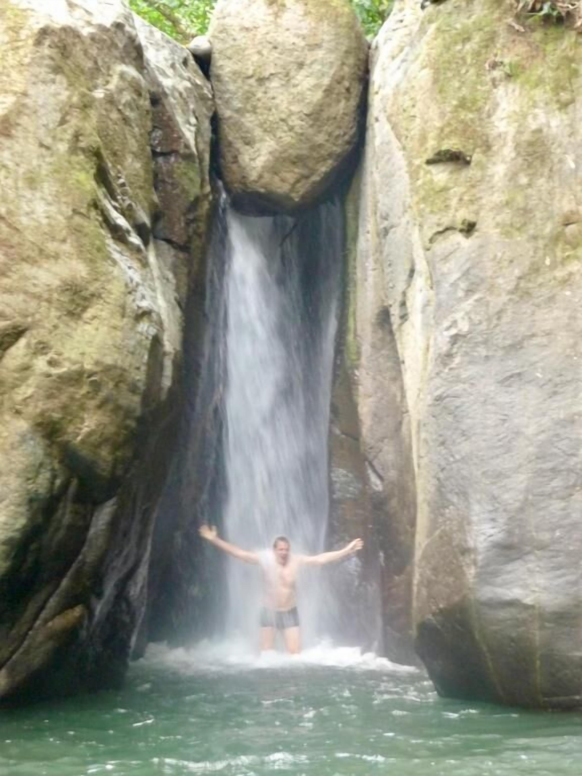 A person standing in a natural water pool with a waterfall behind them.