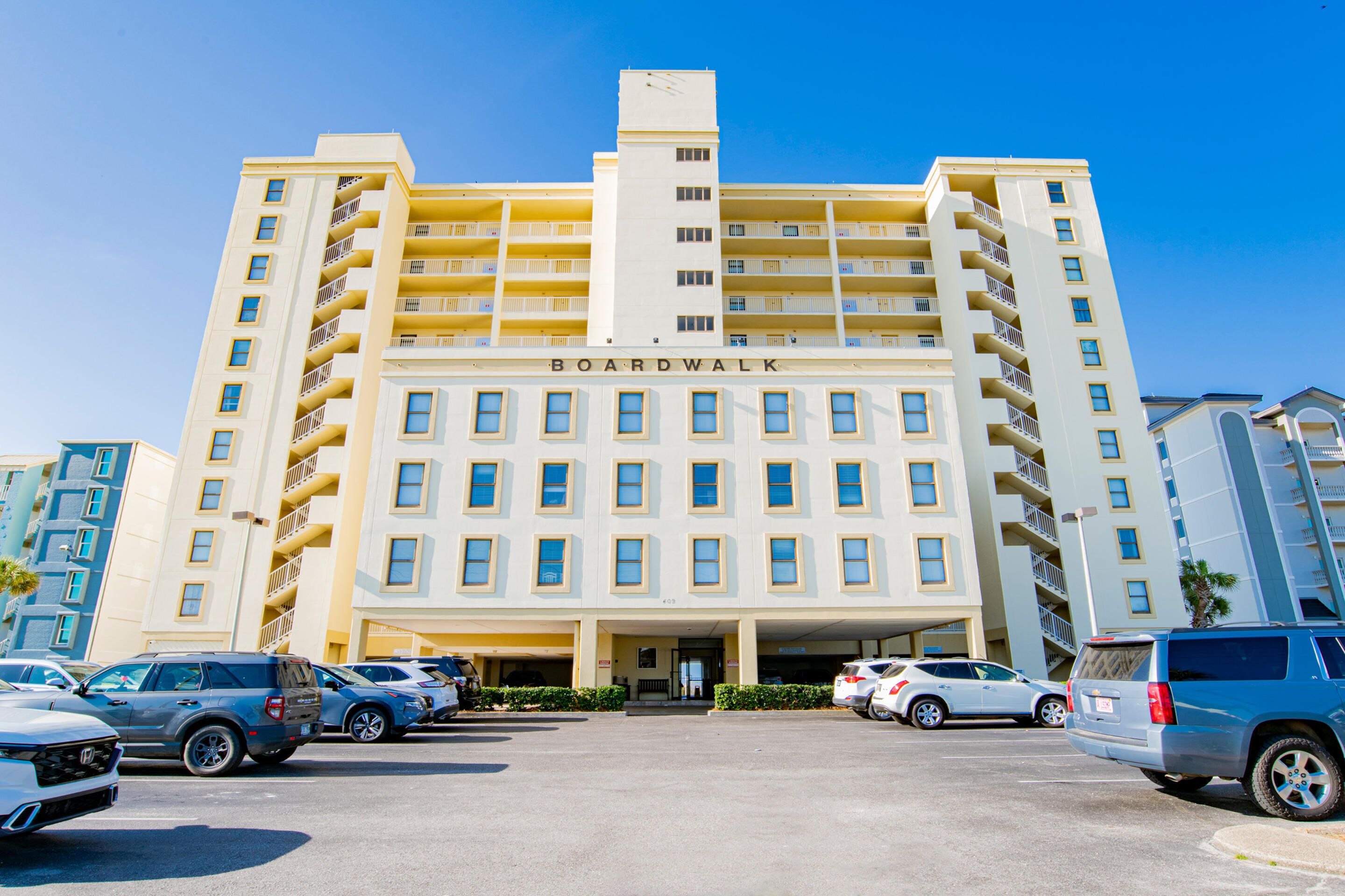 Um hotel com vários andares, entrada central, varandas em cada andar e vários carros estacionados em primeiro plano.