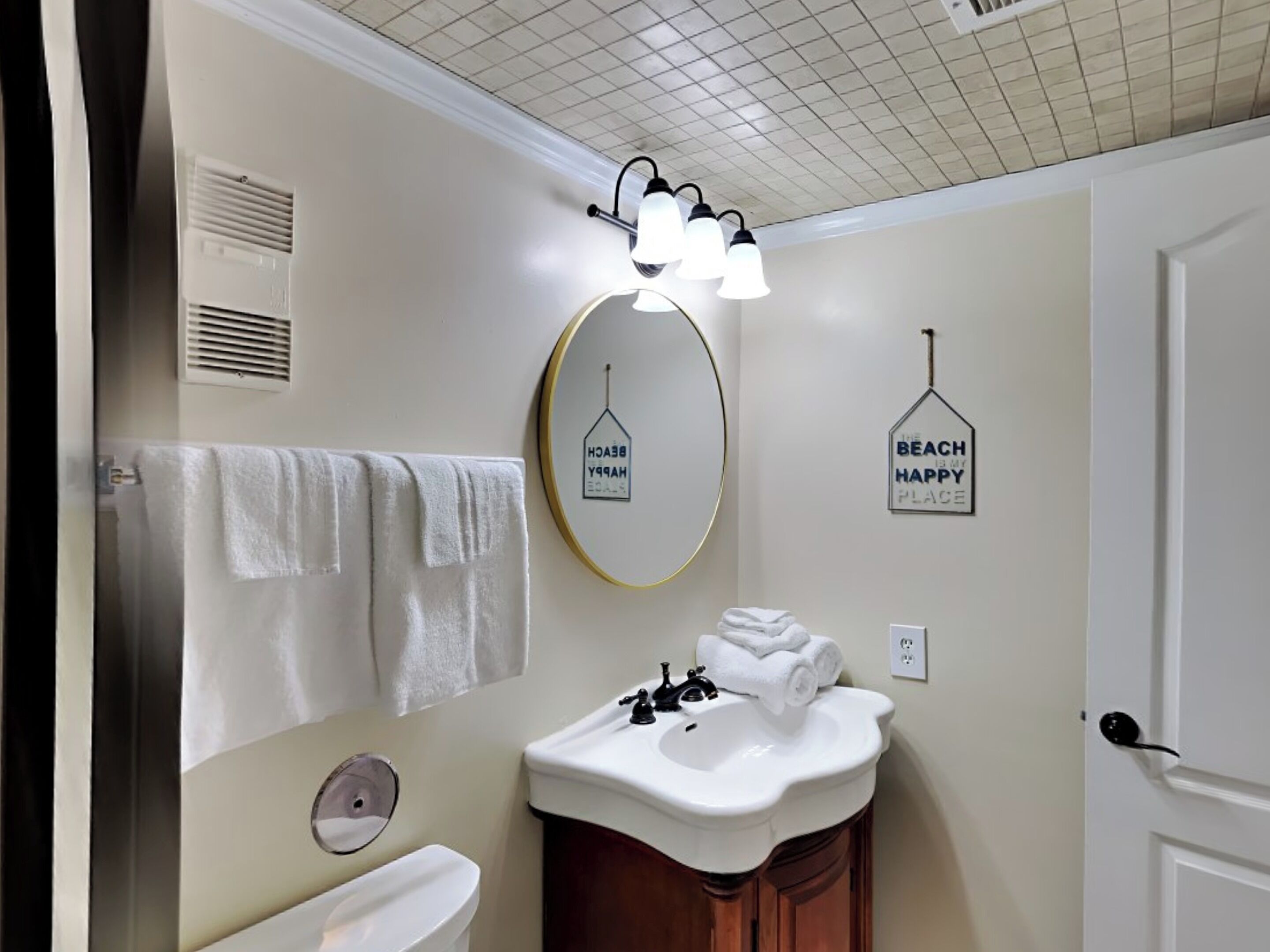 A bathroom with a white sink, a mirror, and a towel rack. There are two wall decorations, one saying "Beach is my happy place" and another with a house icon.