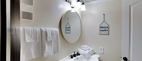 A bathroom with a white sink, a mirror, and a towel rack. There are two wall decorations, one saying "Beach is my happy place" and another with a house icon.