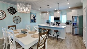 Une cuisine moderne avec des meubles blancs, un îlot central doté de tabourets, un lustre et des œuvres d’art décoratives sur les murs.