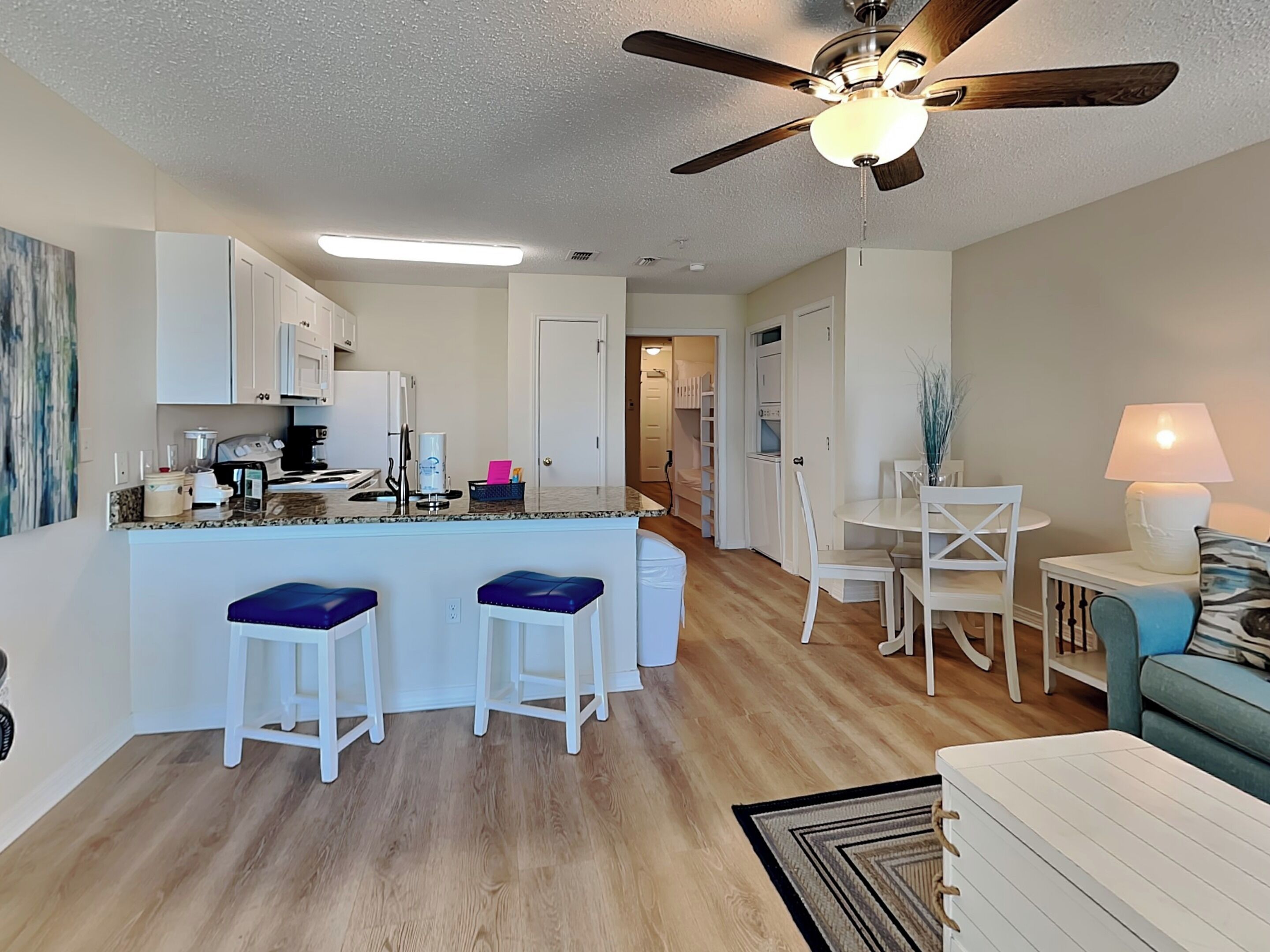 A modern kitchen with white cabinets, a central island with blue bar stools, a ceiling fan, and a dining area with a white table and chairs.
