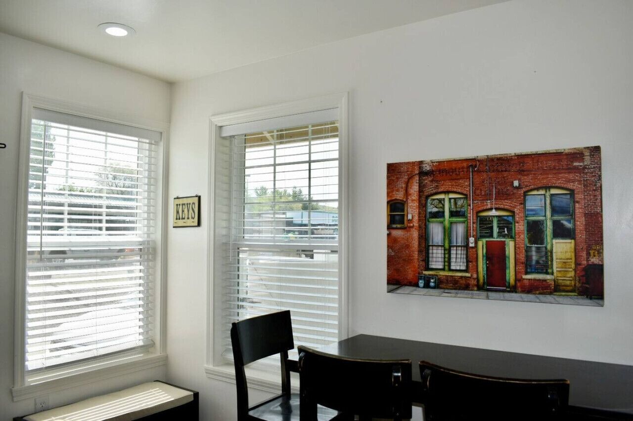 Un coin repas avec une table et des chaises, un tableau représentant un bâtiment accroché au mur et une fenêtre avec des stores.