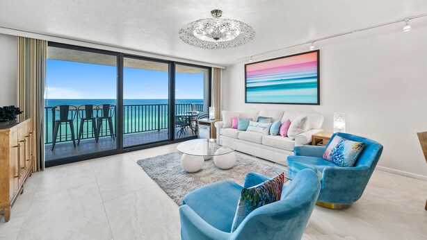 A spacious living room with a large glass door leading to a balcony with a view of the ocean. There is a white sofa, a blue armchair, and a coffee table.