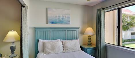 A bedroom with a bed, two bedside lamps, a ceiling fan, a painting, and a window with curtains.