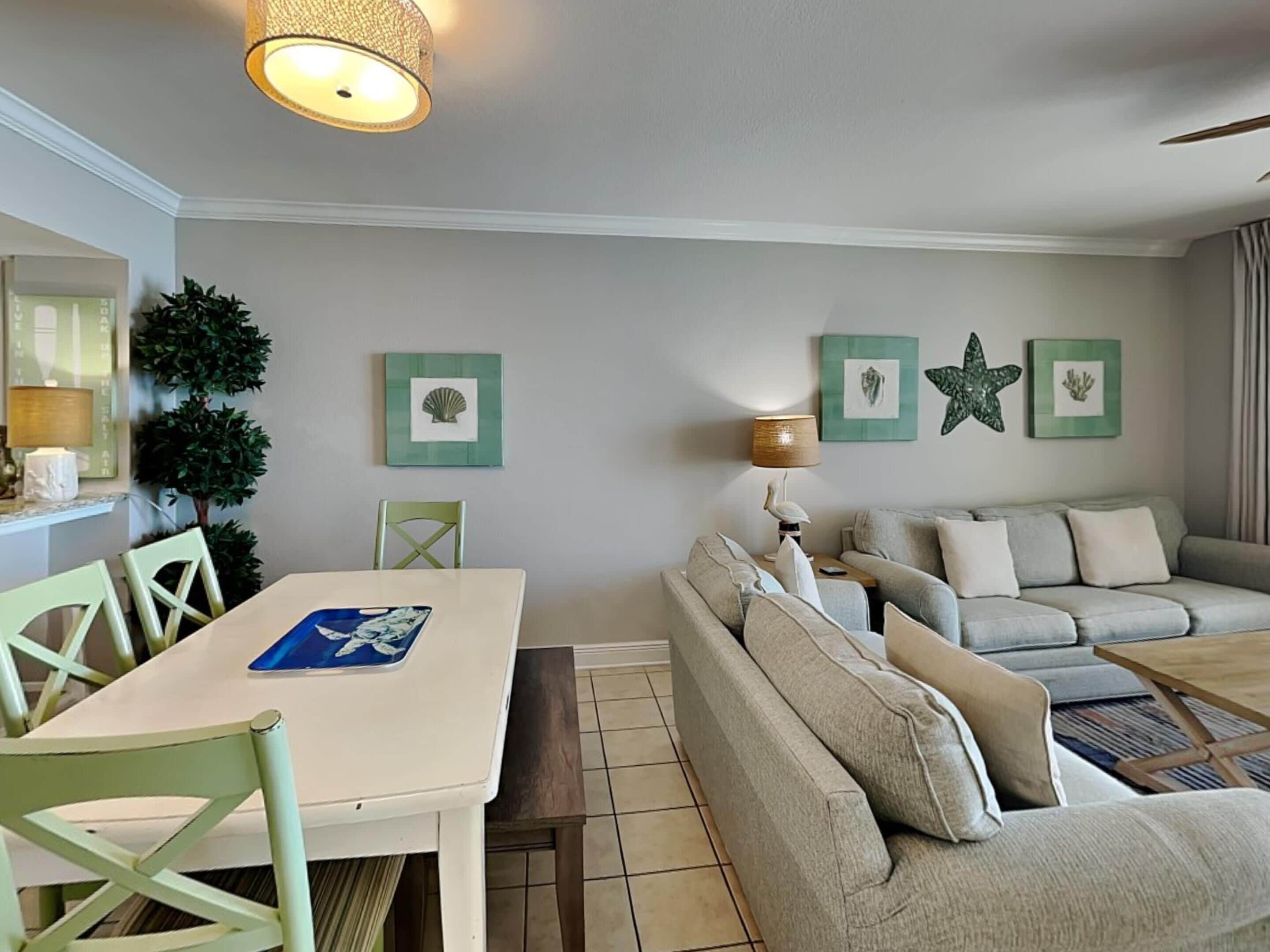 A living room with a dining table, chairs, a sofa, and a lamp. There are framed pictures on the wall and a potted plant.