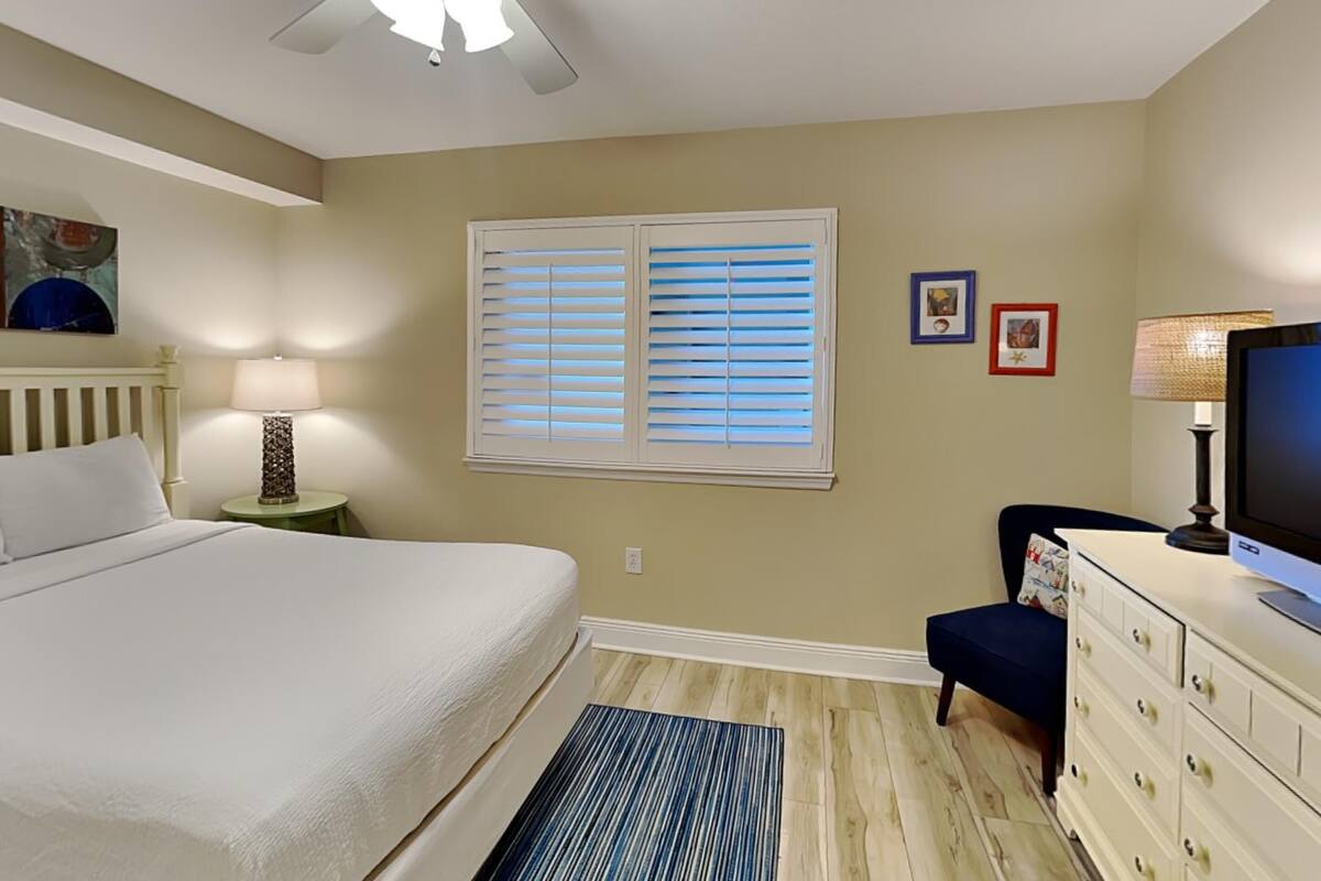 A bedroom with a bed, a ceiling fan, a window with blinds, a desk with a computer, and a dresser.