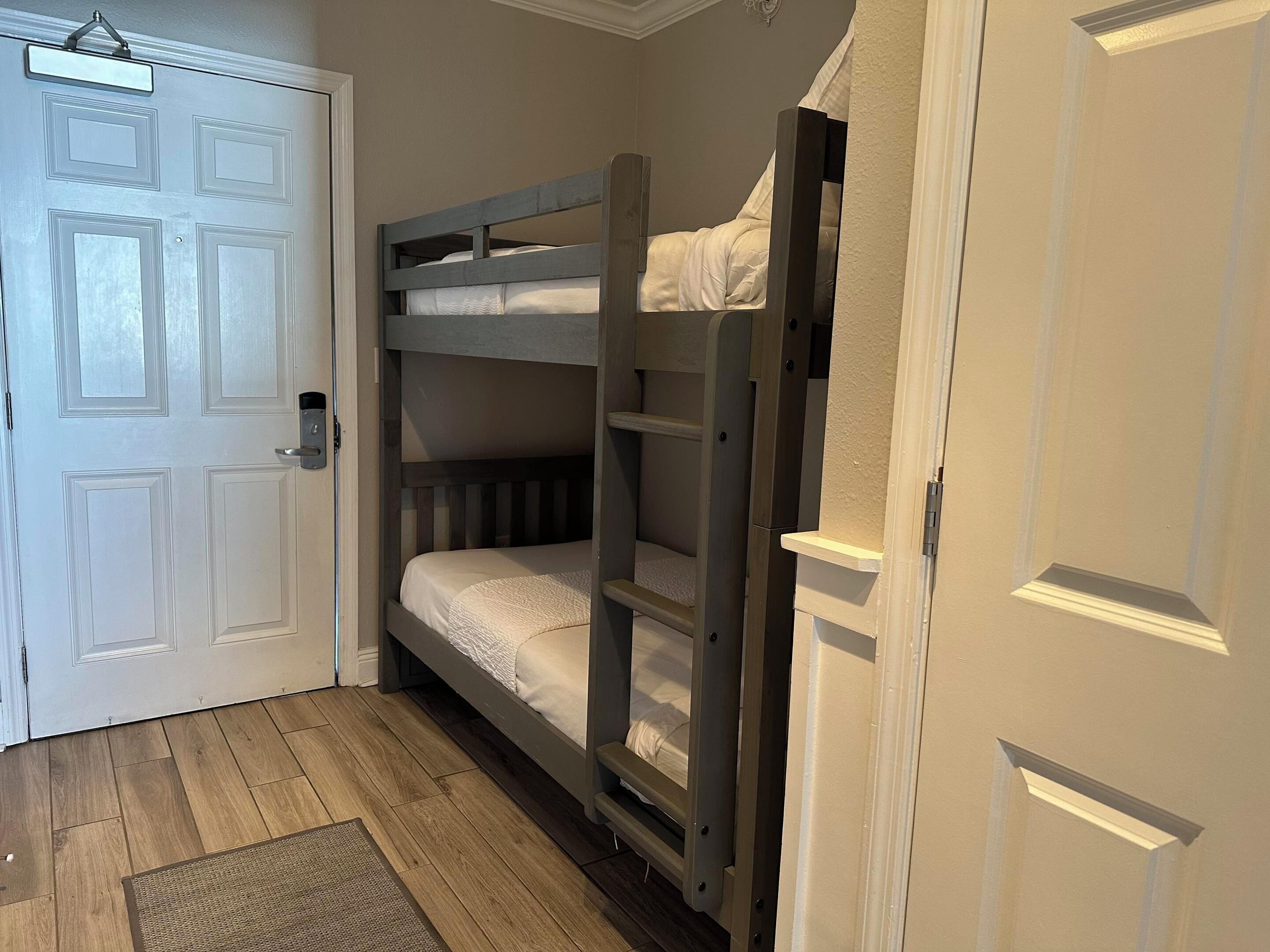 Ein Stockbett mit Holzrahmen und weißer Bettwäsche, eine weiße Tür mit Griff sowie ein Holzboden.
