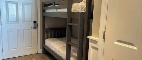 Ein Stockbett mit Holzrahmen und weißer Bettwäsche, eine weiße Tür mit Griff sowie ein Holzboden.