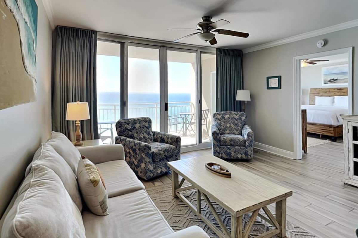A hotel room with a sofa, two patterned armchairs, a coffee table, a ceiling fan, and a view of the ocean through large windows.