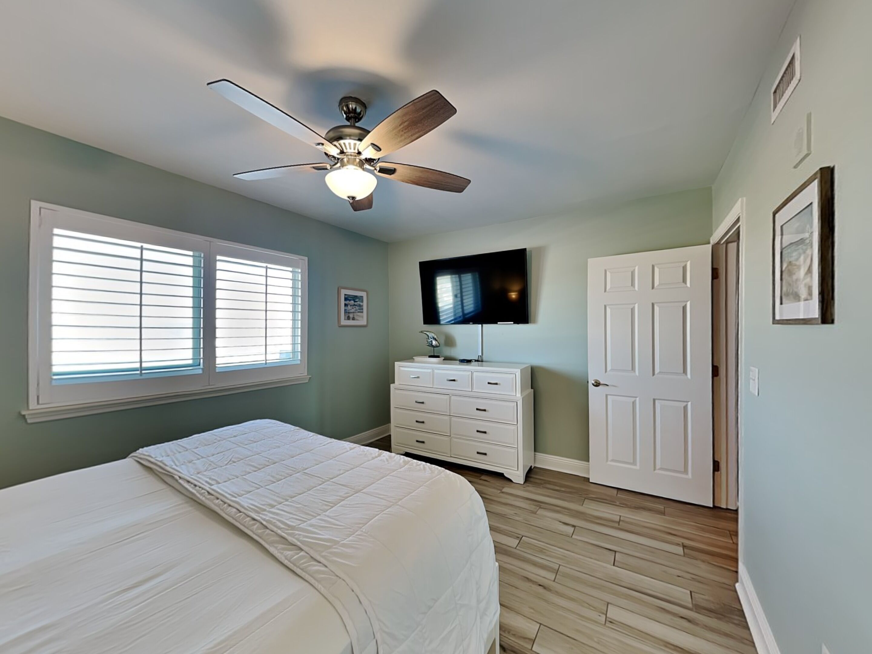 A bedroom with a bed, a ceiling fan, a dresser, a television, and a door.