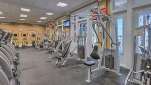 Un gimnasio con diversas máquinas de ejercicio, amplias ventanas y un reloj en la pared.