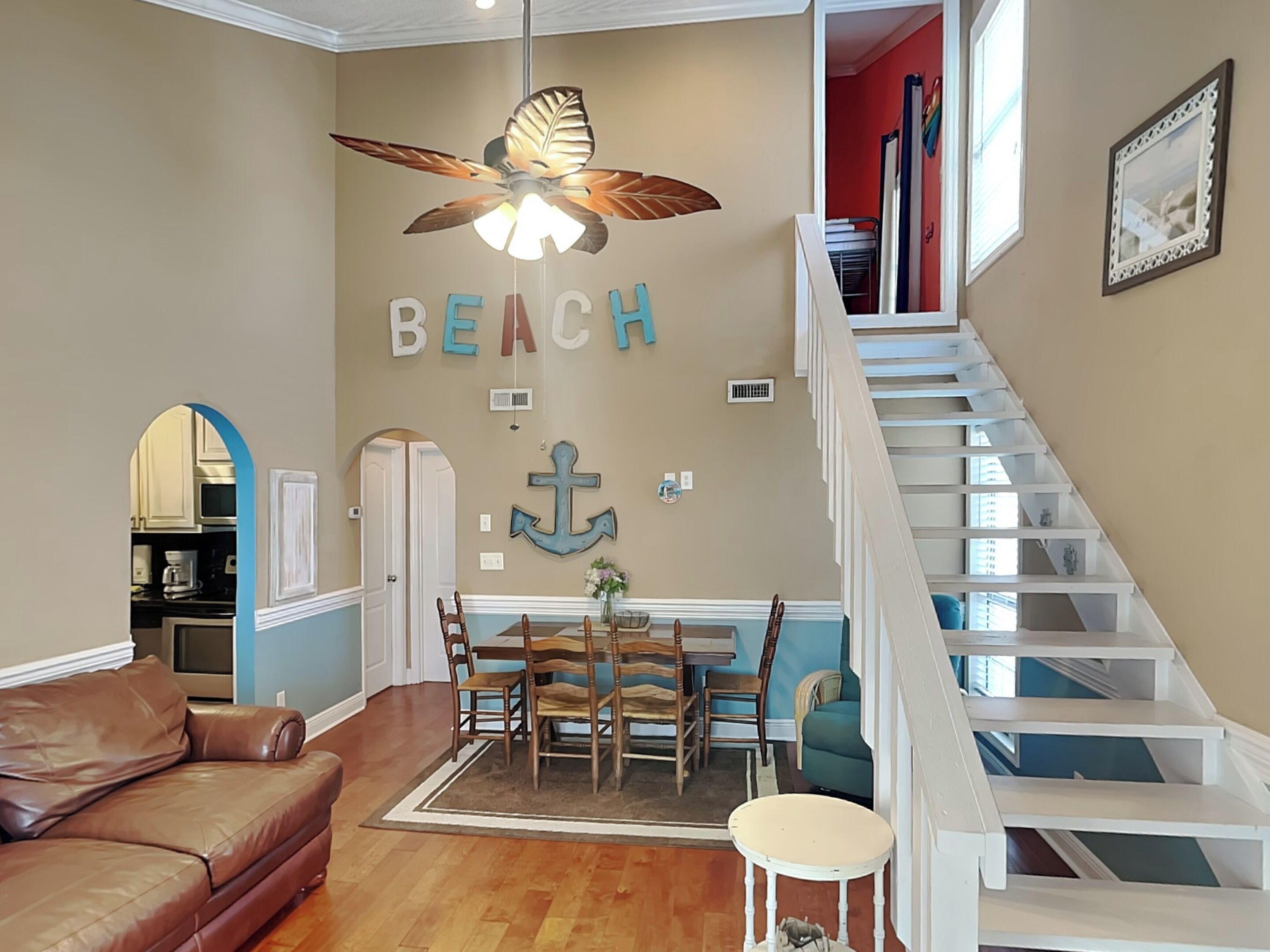 A living room with a beige wall, a ceiling fan, a dining table with chairs, a brown leather couch, and a staircase with white railings.