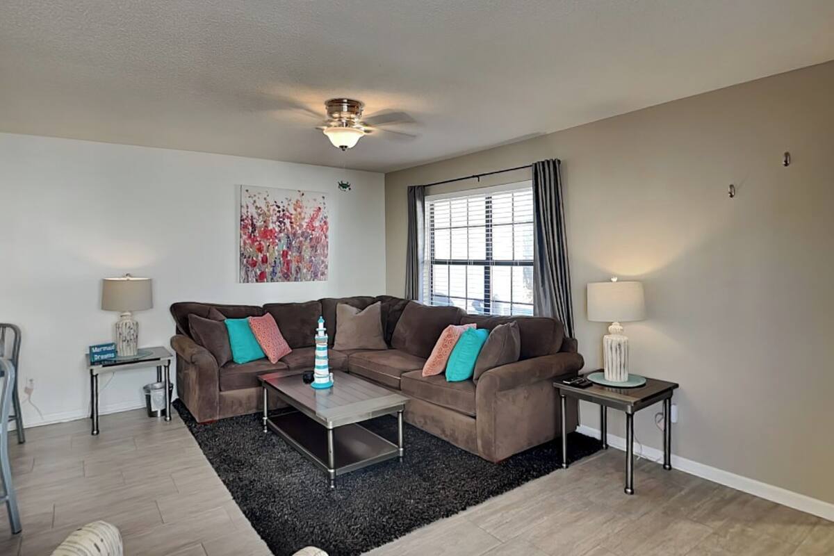 A living room with a brown sectional sofa, a coffee table, a side table with a lamp, a window with curtains, and a ceiling fan.