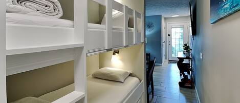 A hotel room with a bunk bed, a desk, and a chair. There is a door leading to a hallway with a dining area.