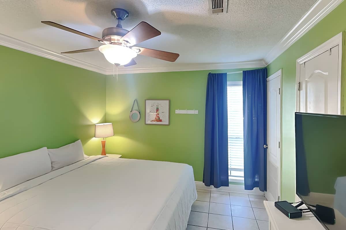 A hotel room with a bed, a ceiling fan, a nightstand with a lamp, a picture on the wall, and a window with curtains.