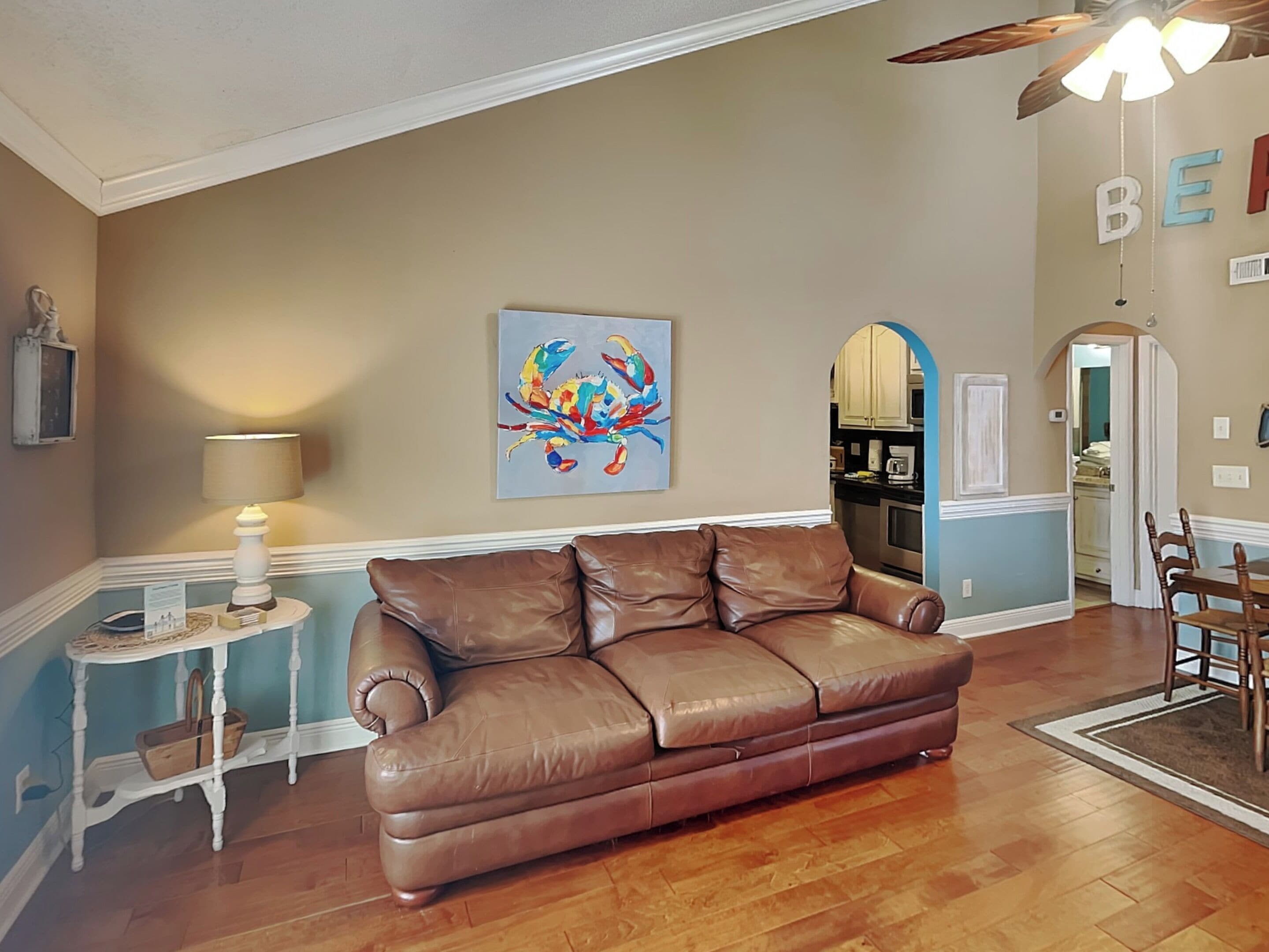 A living room with a brown leather sofa, a small side table with a lamp, a colorful abstract painting, and a kitchen visible through an archway.