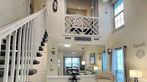 Un vaste espace de vie doté d’un haut plafond, d’un escalier avec des rampes blanches, d’un canapé en cuir beige et d’un coin repas agrémenté d’un lustre.
