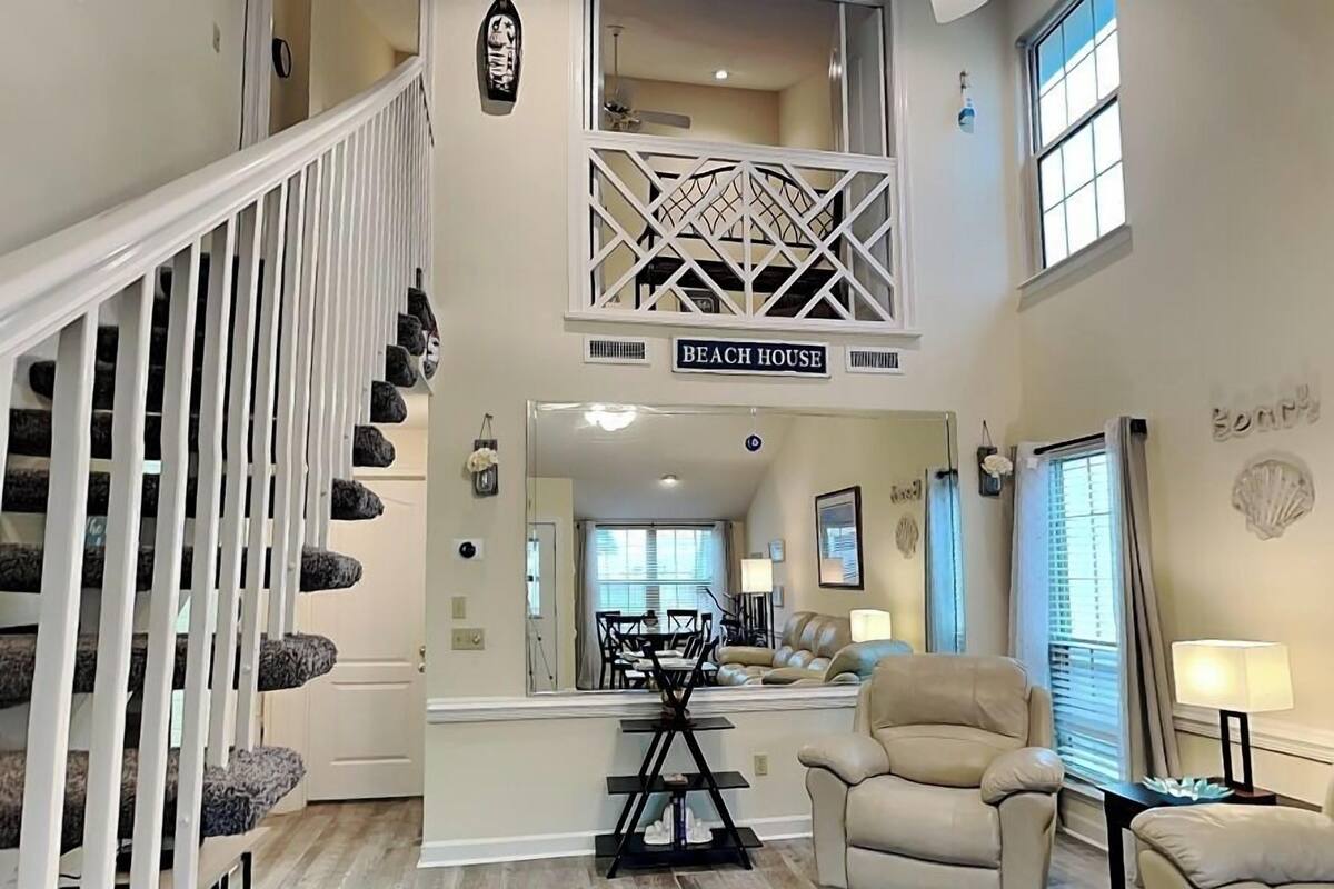 A spacious living area with a high ceiling, a staircase with white railings, a beige leather sofa, and a dining area with a chandelier.