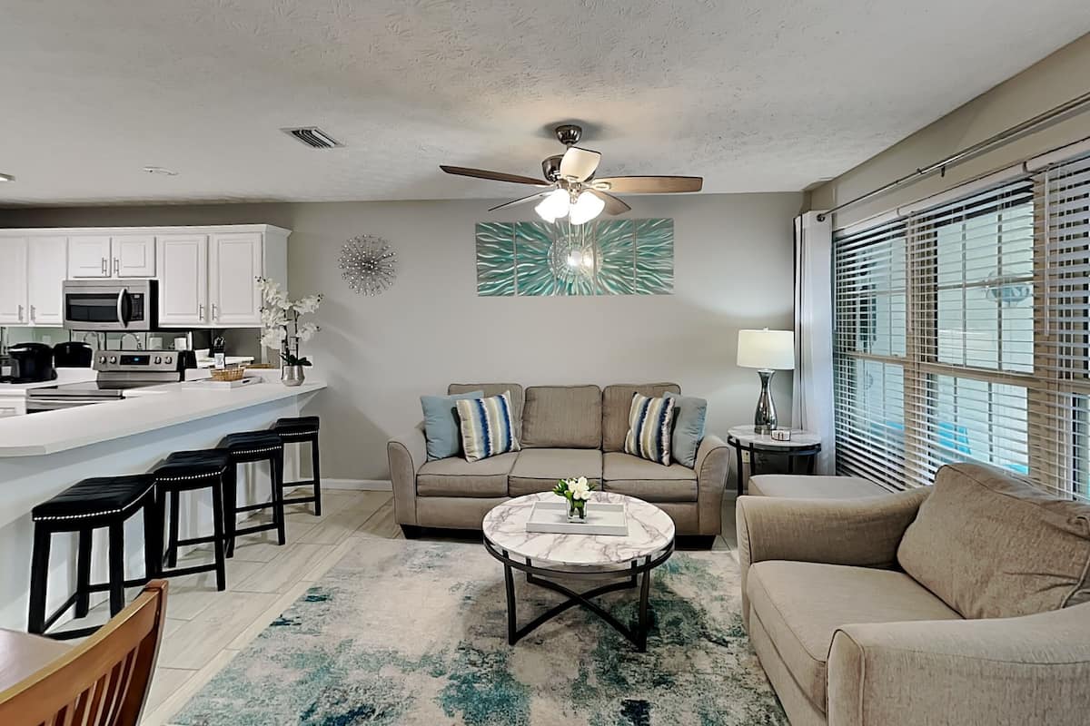 A modern living room with a sofa, coffee table, and chairs. There is a kitchen area with appliances and a dining table.