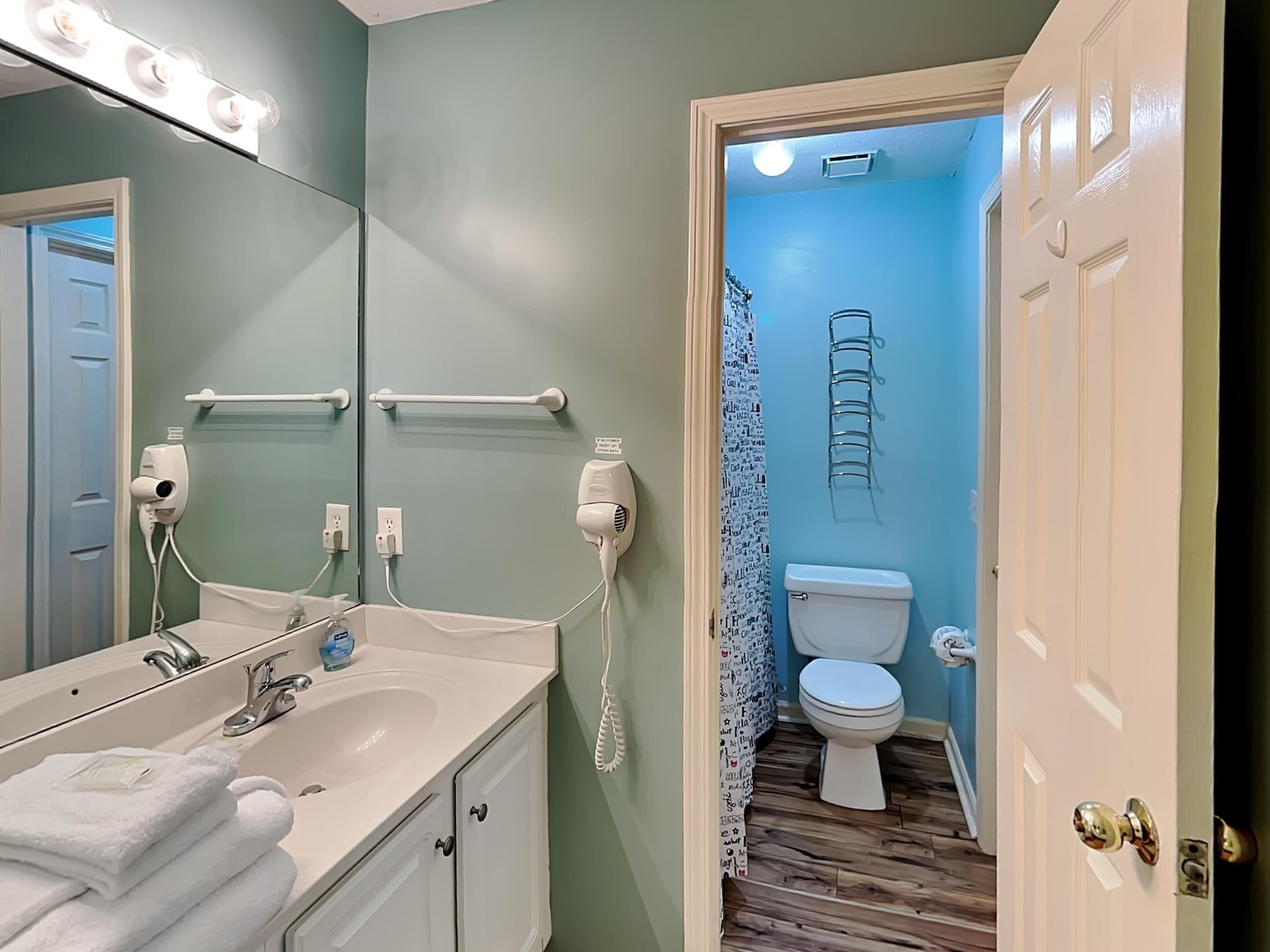 A bathroom with a vanity, a sink, a mirror with lights, a hairdryer, a toilet, and a shower curtain.