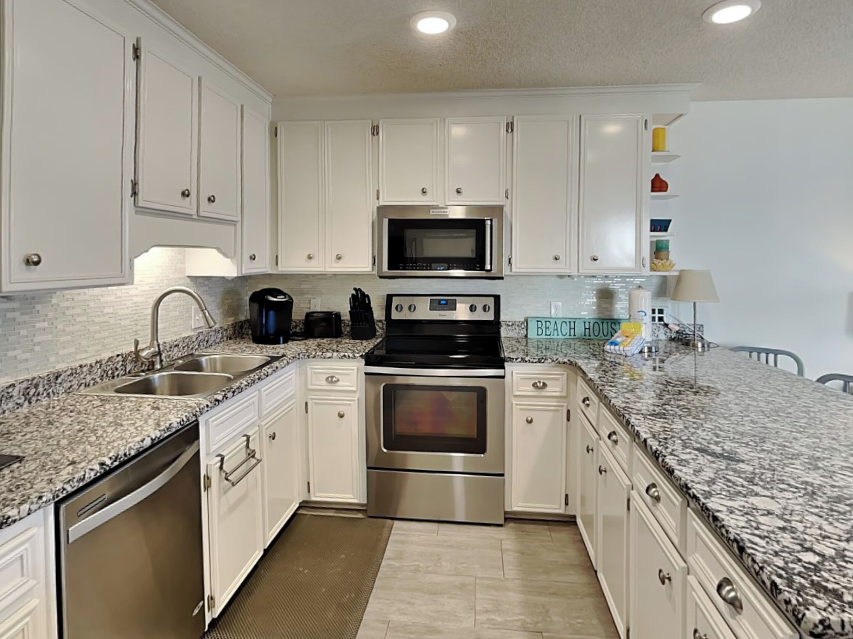 A modern kitchen with white cabinets, stainless steel appliances, granite countertops, and a sign reading "BEACH HOUSE".