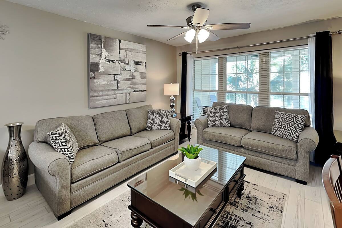 A living room with two grey sofas, a coffee table, a floor lamp, a ceiling fan, and a large abstract painting on the wall.