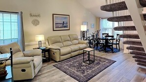 Un salon avec un canapé beige, une table basse, un tapis et un coin repas comprenant des chaises et une table. Un escalier se trouve sur la droite, avec un mur à texture.