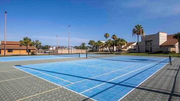 An outdoor tennis court with a net, surrounded by palm trees and buildings.