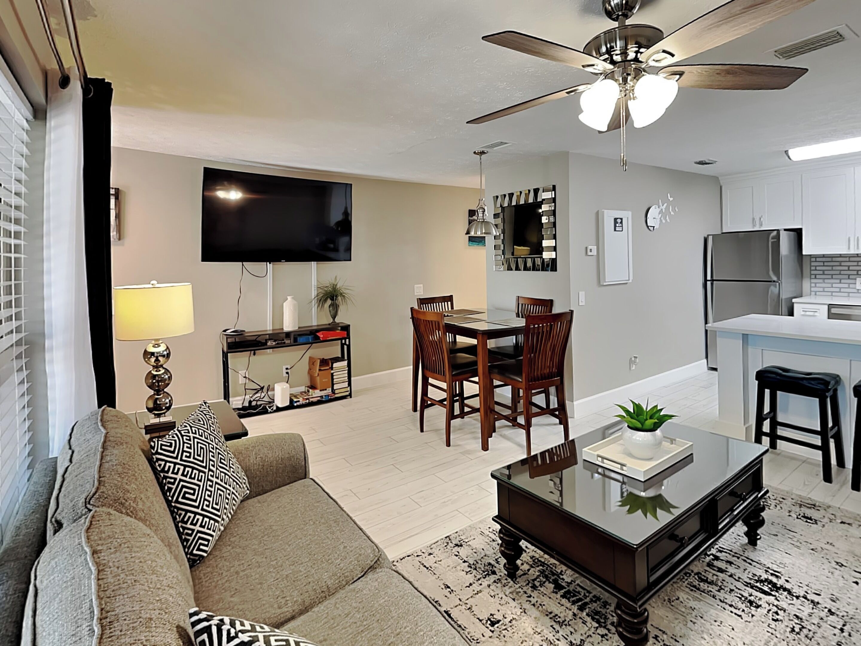 A modern living room with a sofa, coffee table, and dining area. There is a ceiling fan, a flat-screen TV, and a kitchen with a refrigerator.