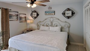 Un dormitorio con una cama grande, un ventilador de techo y decoración en la pared, incluyendo una escena de playa y un pájaro.