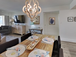 A dining area with a wooden table set for four, surrounded by dark chairs. A chandelier hangs above the table, and a television is mounted on the wall to the left.