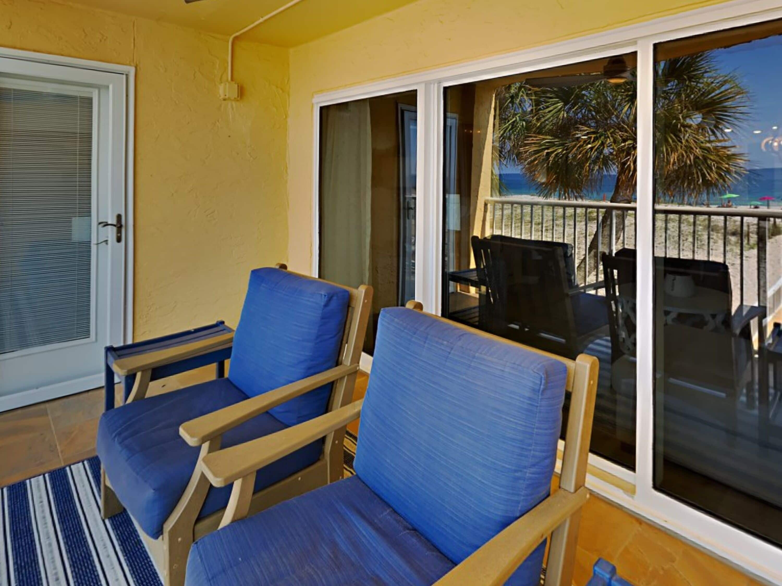 A room with two blue cushioned chairs, a wooden table, and a large sliding glass door leading to a balcony with a view of palm trees and a beach.
