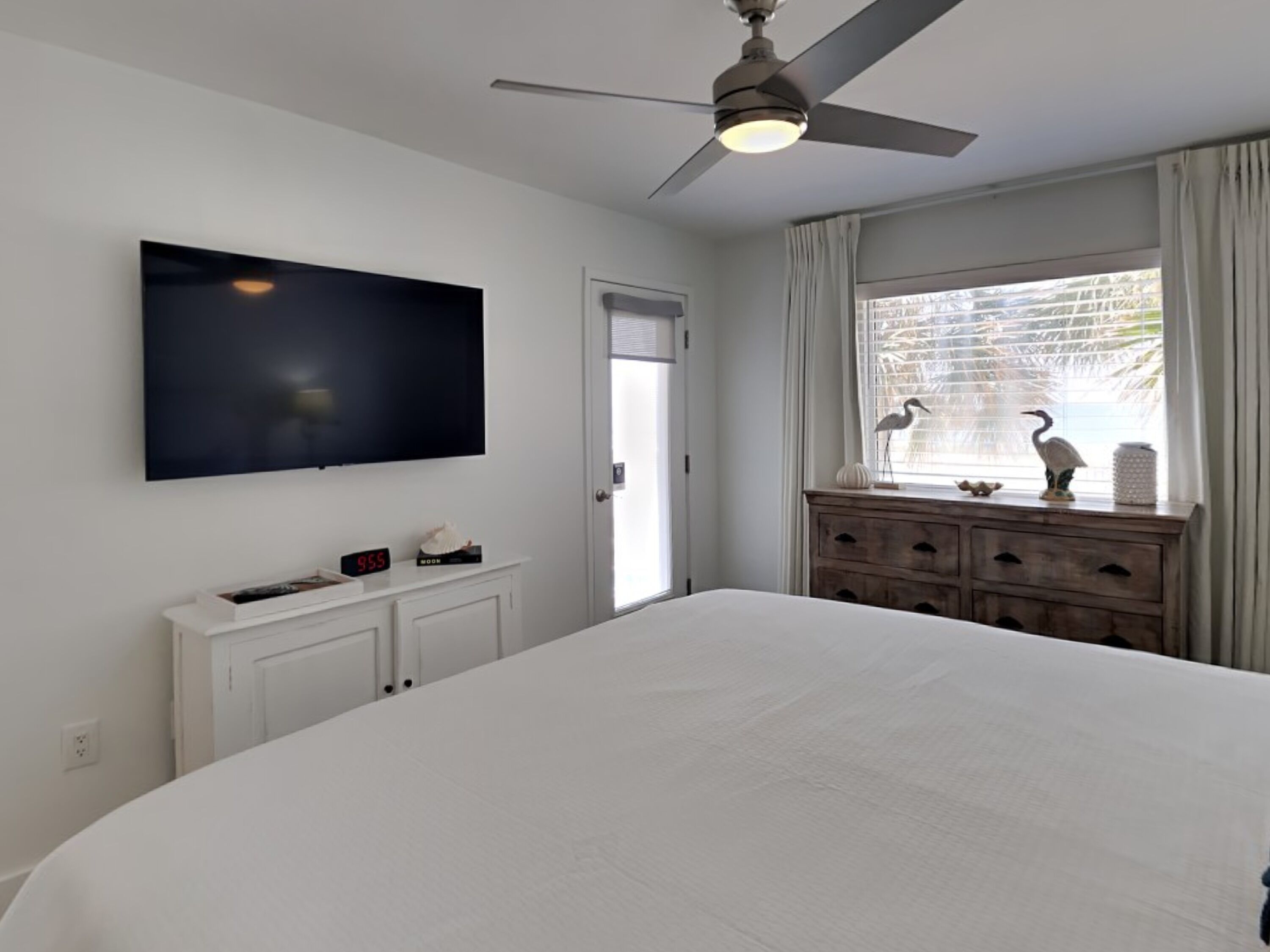 Un dormitorio con una cama grande, un ventilador de techo, un televisor montado en la pared, un aparador con objetos decorativos y una ventana con cortinas.