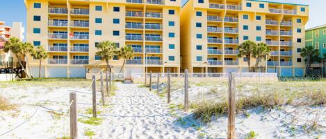A multi-story hotel with balconies, located near a sandy beach area.
