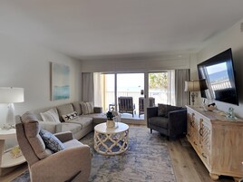 Ein geräumiges Wohnzimmer mit einer großen Couch, einem kleinen Sessel, einem Couchtisch und einem an der Wand montierten Fernseher.