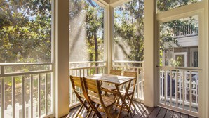 A covered porch with a wooden table and chairs, surrounded by white railings and large windows offering views of trees and a building. - Anchored on 30A by Southern Vacation Rentals (Santa Rosa Beach)
