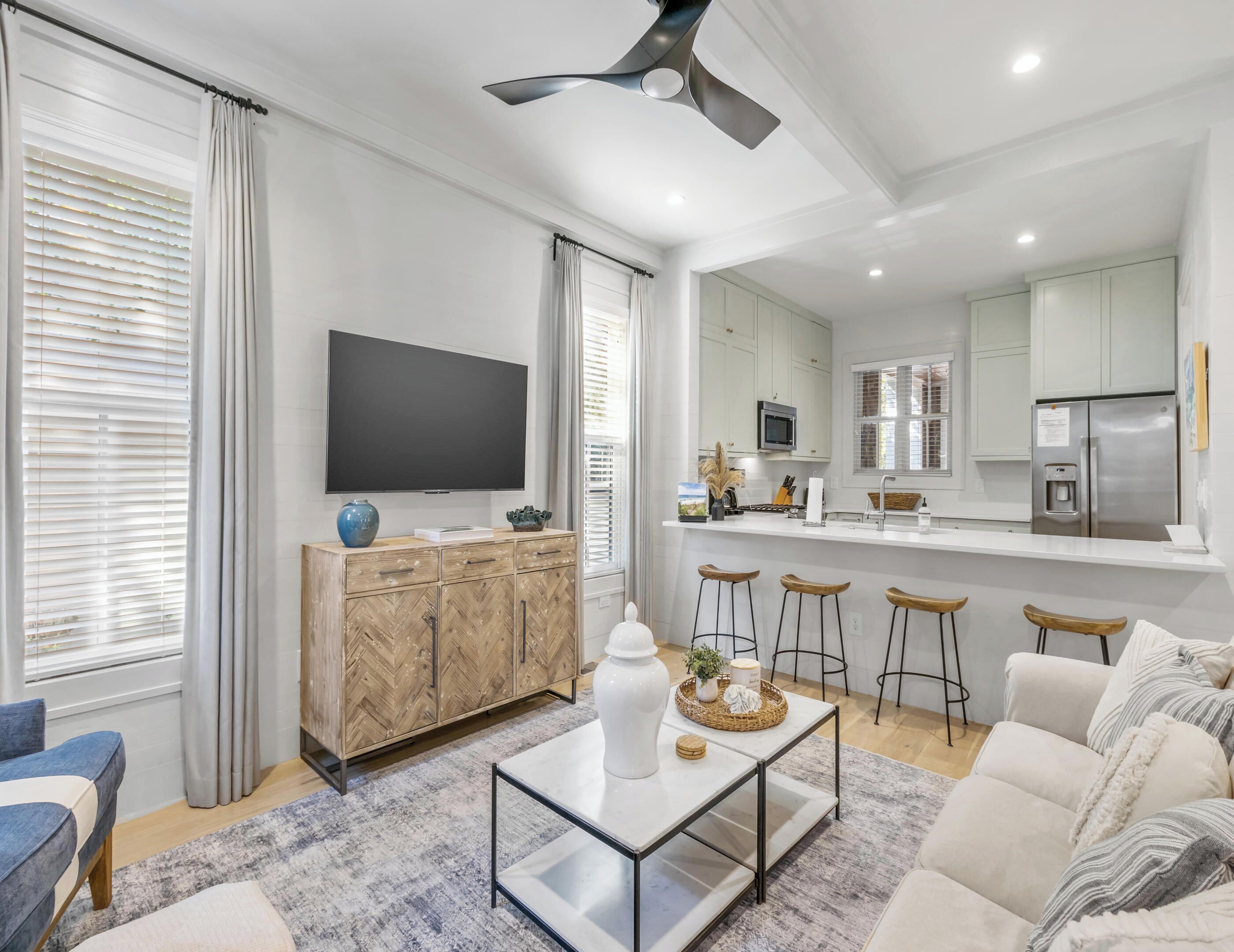 A modern living room with a sofa, a coffee table, and a TV. There is a kitchen area with a fridge, microwave, and bar stools.