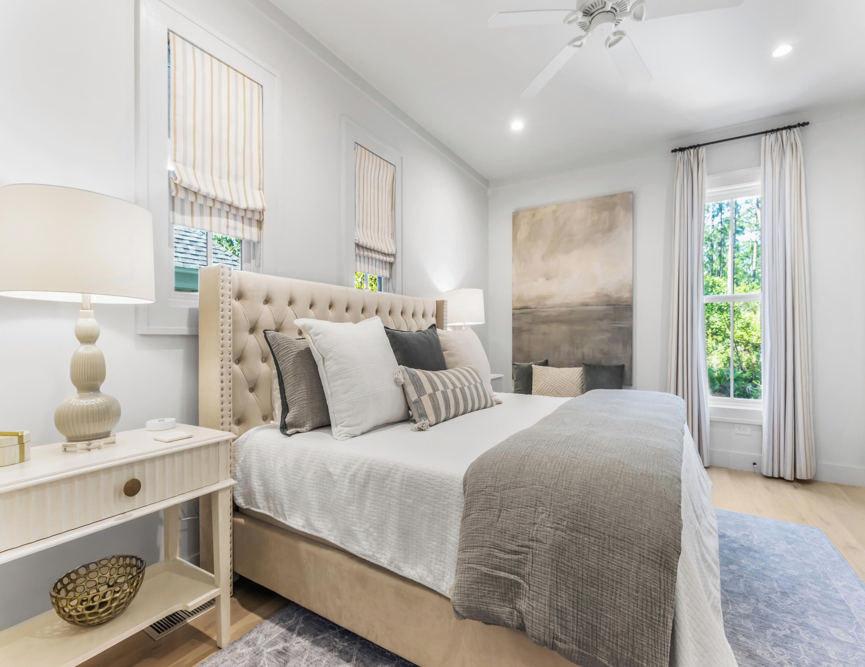 A modern bedroom with a tufted headboard, a large bed with multiple pillows, a bedside table with a lamp, and a view of greenery through the window.