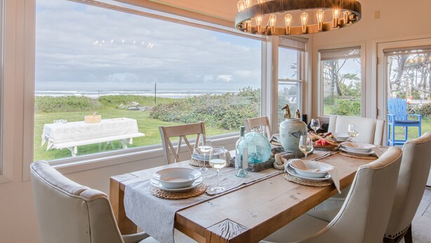 Uma área de refeições com vista para o oceano, uma mesa de madeira posta para quatro pessoas e um lustre moderno.