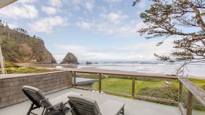 Um pátio com duas poltronas de descanso e uma mesa, com vista para uma praia, um penhasco rochoso e o oceano.