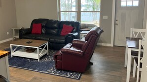 A living room with a black leather sofa, a red leather chair, a coffee table, and a rug. There is a window with a view of trees and a door.