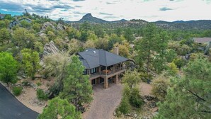 A modern house with a large deck, surrounded by dense forest and rocky terrain. - Mountain Retreat | Hot Tub & Fire Pit (Prescott)