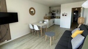 A modern living room with a black sofa, a dining table with four chairs, a kitchen area with white cabinets, and a TV mounted on the wall. - Seignosse Ocean, beautiful renovated apartment OCEAN VIEW 150m from the beach, ideal family (SEIGNOSSE)