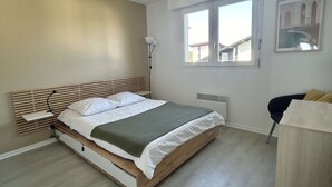 A bedroom with a bed, bedside table, lamp, radiator, window, and a chair. - Seignosse Ocean, beautiful renovated apartment OCEAN VIEW 150m from the beach, ideal family (SEIGNOSSE)
