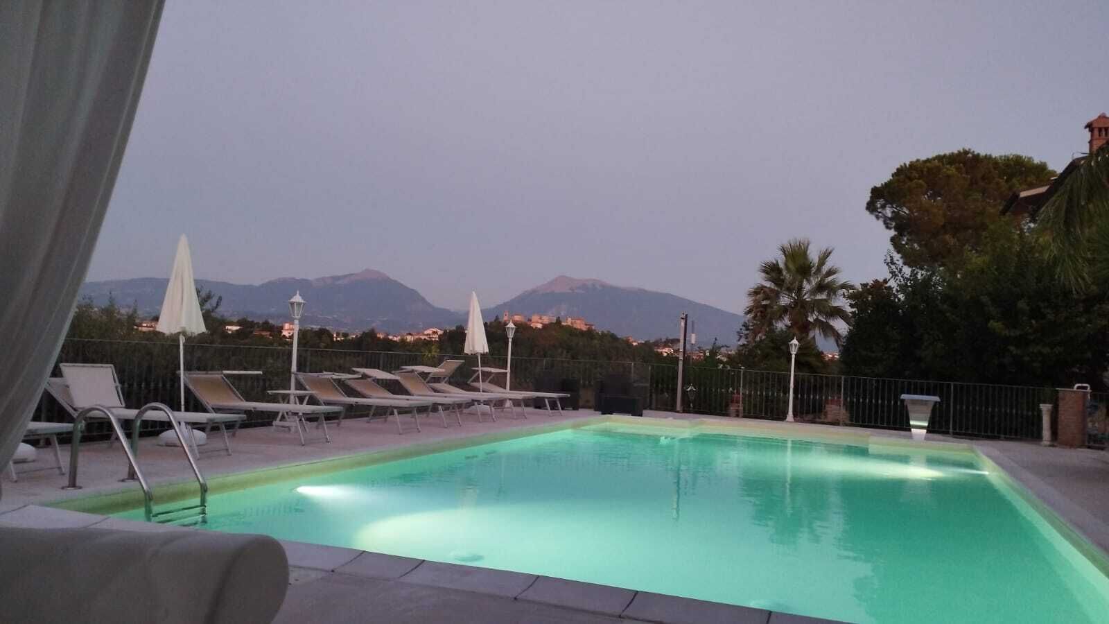 Piscina com espreguiçadeiras e guarda-sóis, cercada por uma grade, com montanhas ao fundo.