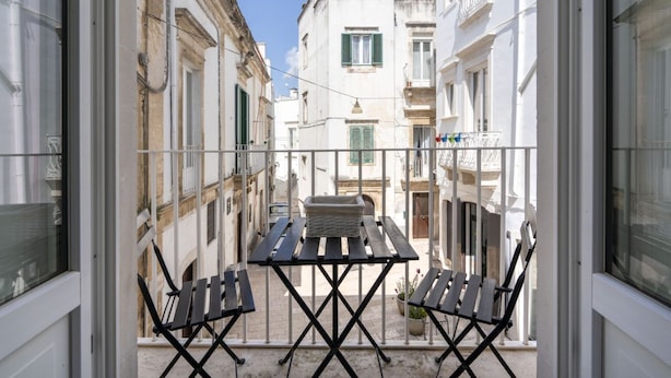 A balcony with a table and chairs, overlooking a narrow street with buildings.